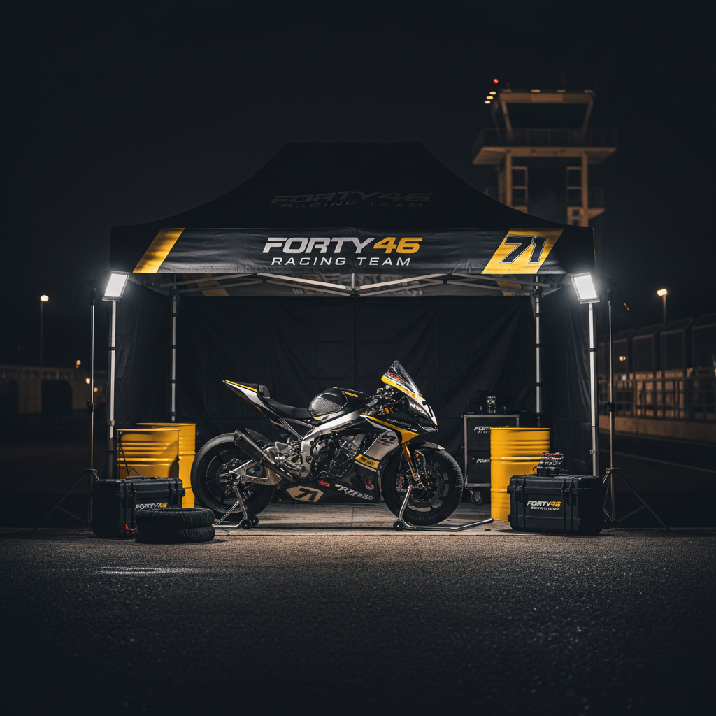 A night-time paddock scene featuring the closed Forty Six Racing Team awning, its black and yellow branding and number 71 graphics glowing under powerful portable LED floodlights. In front of the awning, the team’s race bike sits on stands, spotless and fully prepared, its polished metal and glossy paint catching dramatic rim lighting against the dark surroundings. Fuel drums, tire warmers, and labeled equipment cases sit orderly around the bike, while the racetrack control tower and distant pit lane lights form a soft bokeh in the background. Captured in photographic realism from a slightly elevated angle, the composition uses strong contrasts between bright highlights and deep shadows to create a bold, intense, almost cinematic atmosphere, suggesting readiness for an important race day ahead.