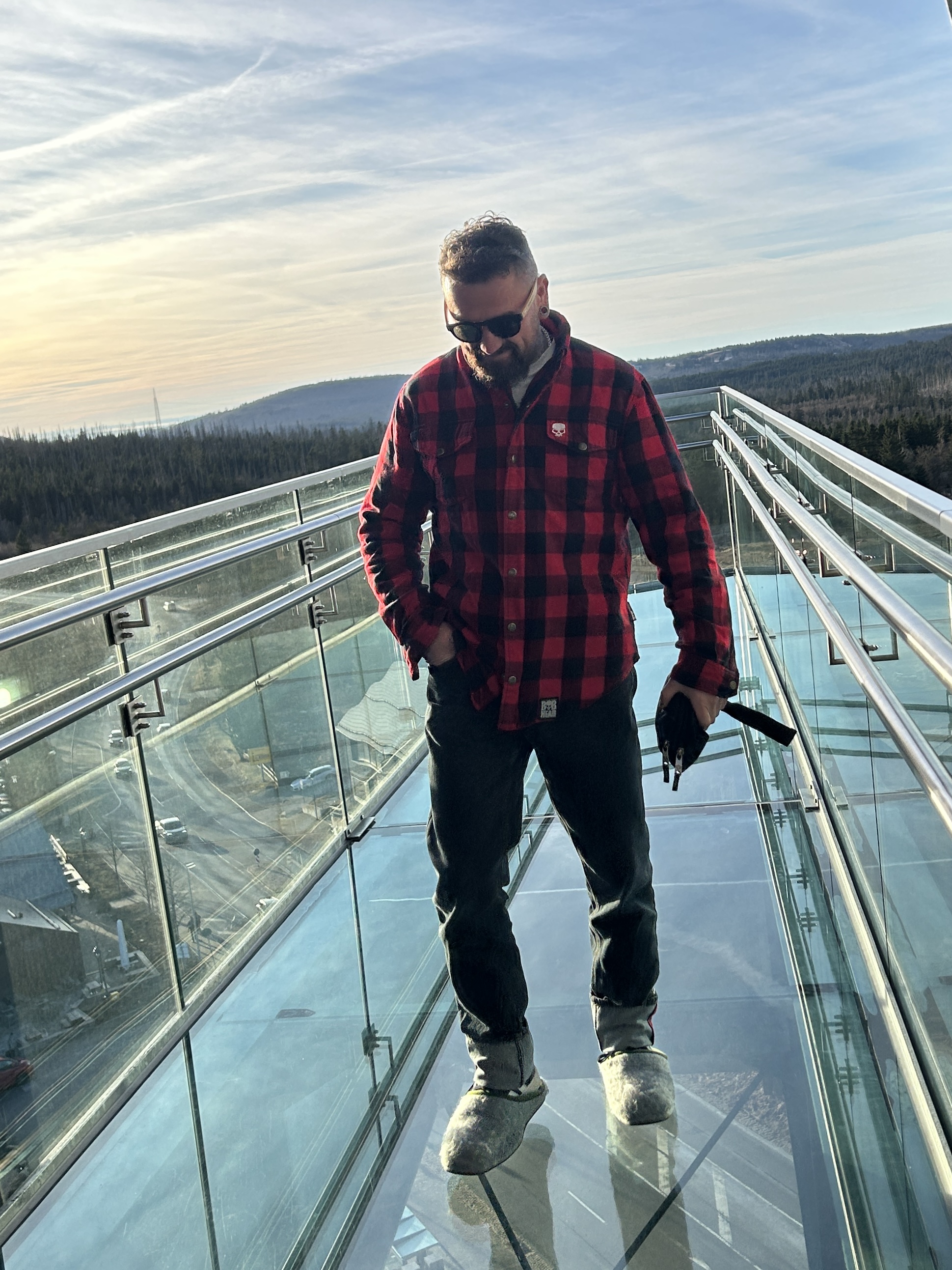 Ein Mann in rotem Karohemd und Sonnenbrille steht auf einem Glassichtboden mit Aussicht auf Wälder und Berge im Hintergrund